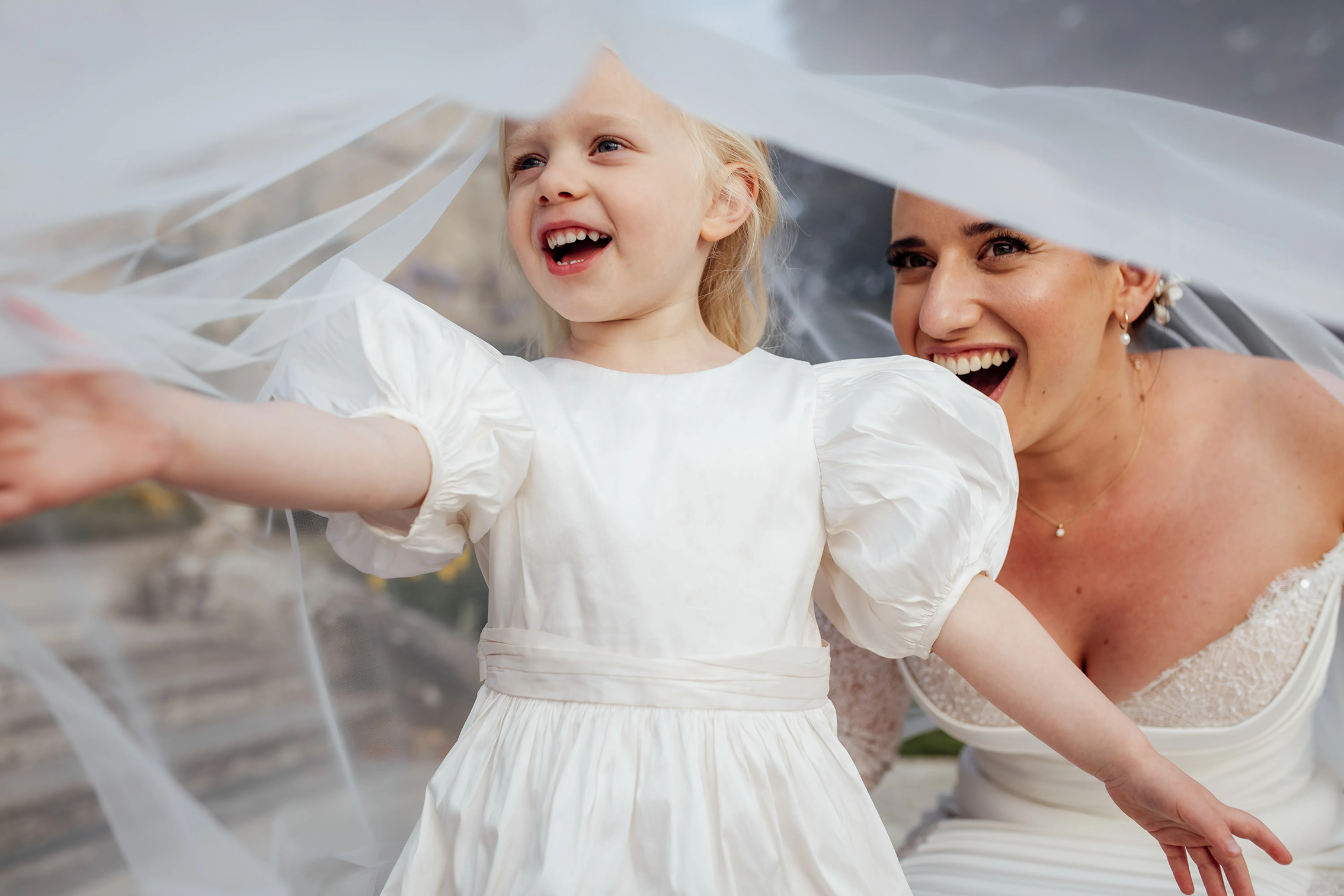 A young girl in a white dress joyfully reaches out with her arms, surrounded by flowing wedding veils. A bride sits nearby