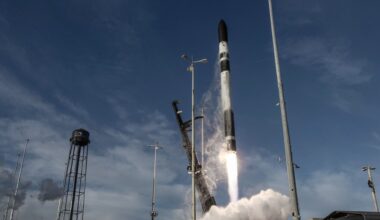a black and white rocket lifts off above a cone of fire and smoke next two a black water tower