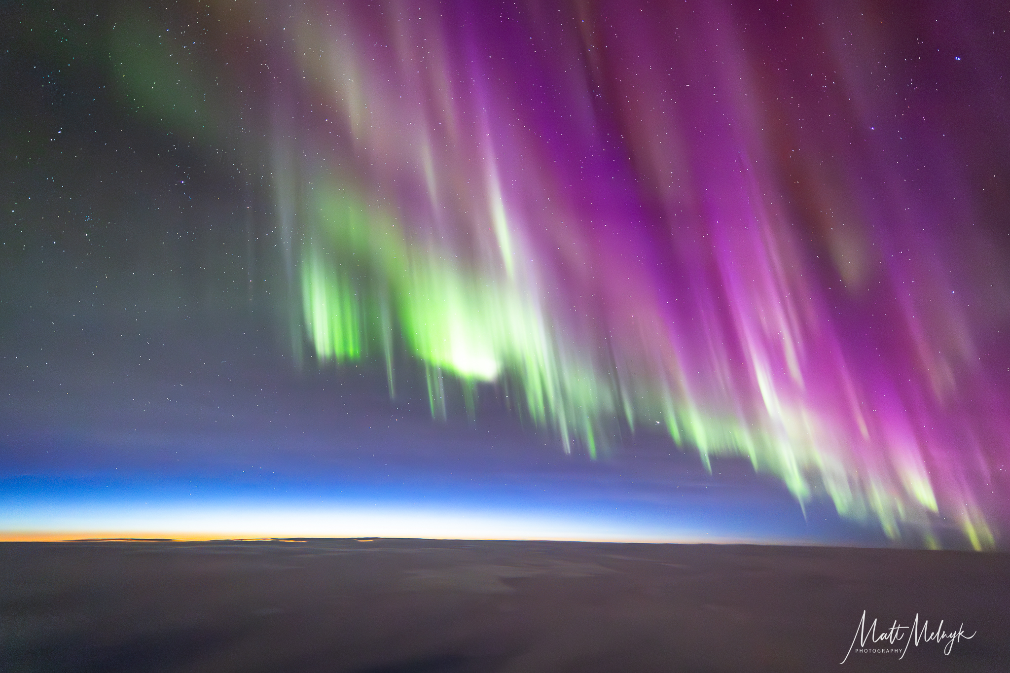 curtains of magenta and green light dance in the sky, a thin blue line marking Earth's atmosphere is visible in the distance. 