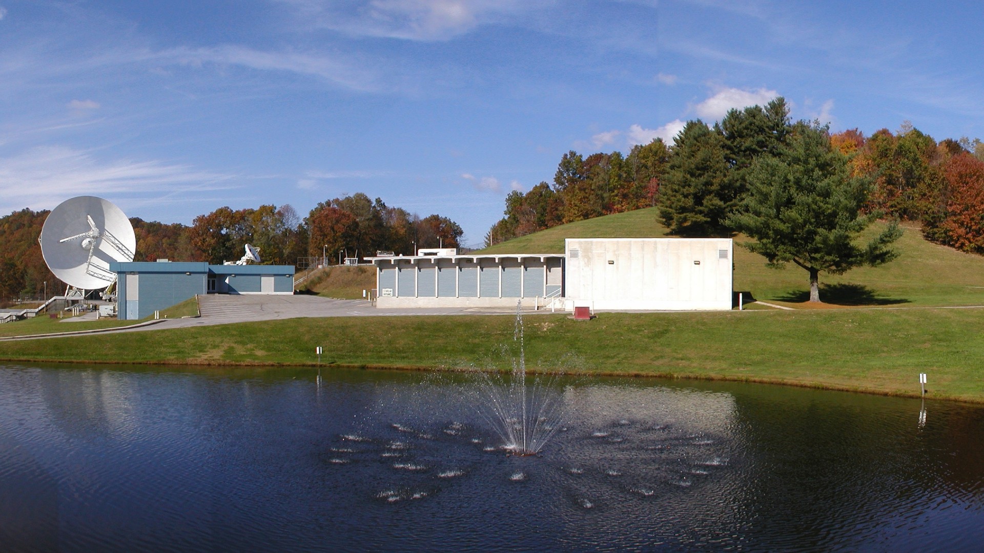 a lake with a fountain in front of several concrete buildings and a large radio telescope dish