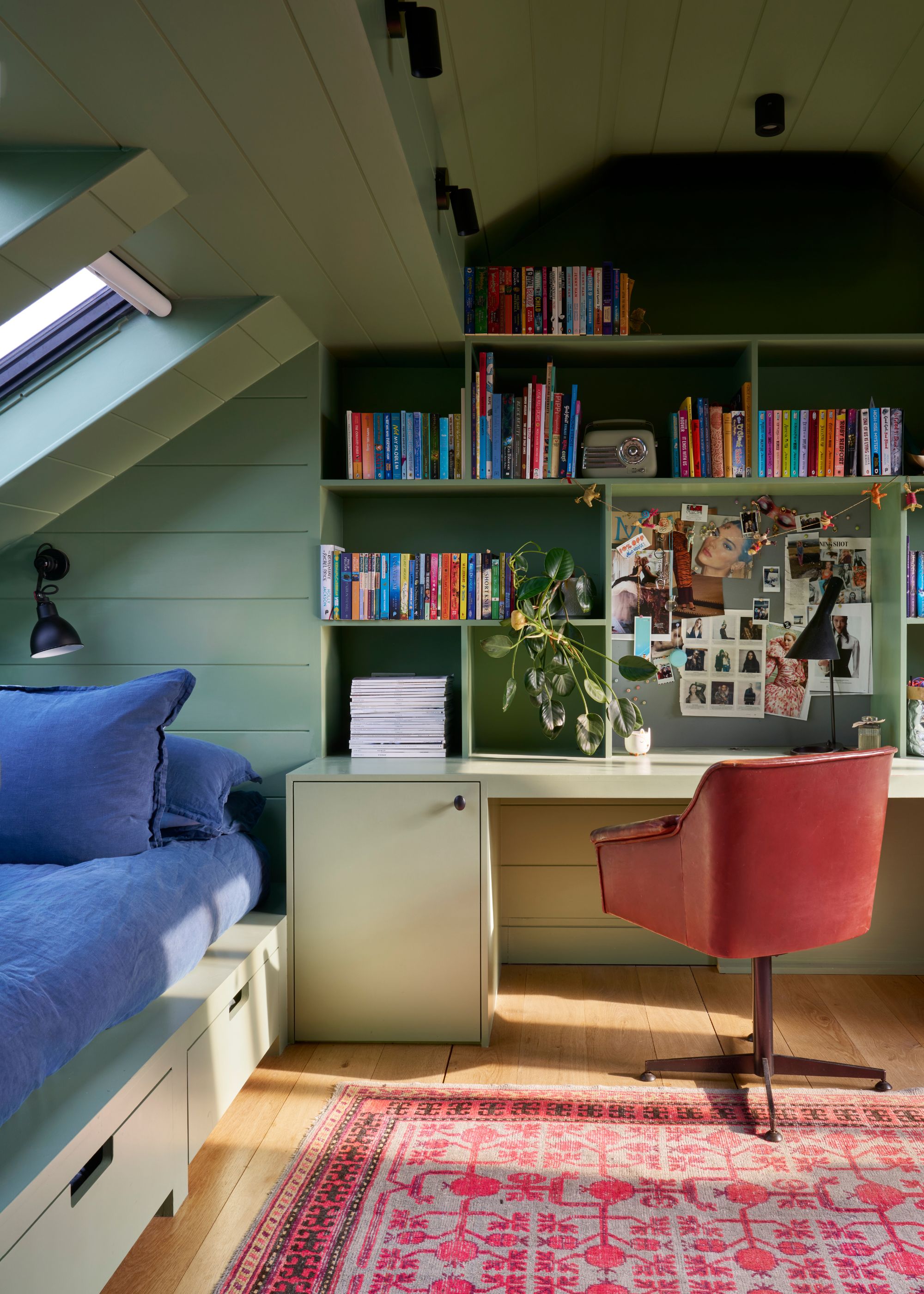 A bedroom painted green with book storage as well as a bench beside the window. There is also a desk below the book shelf with an arm chair next to it.