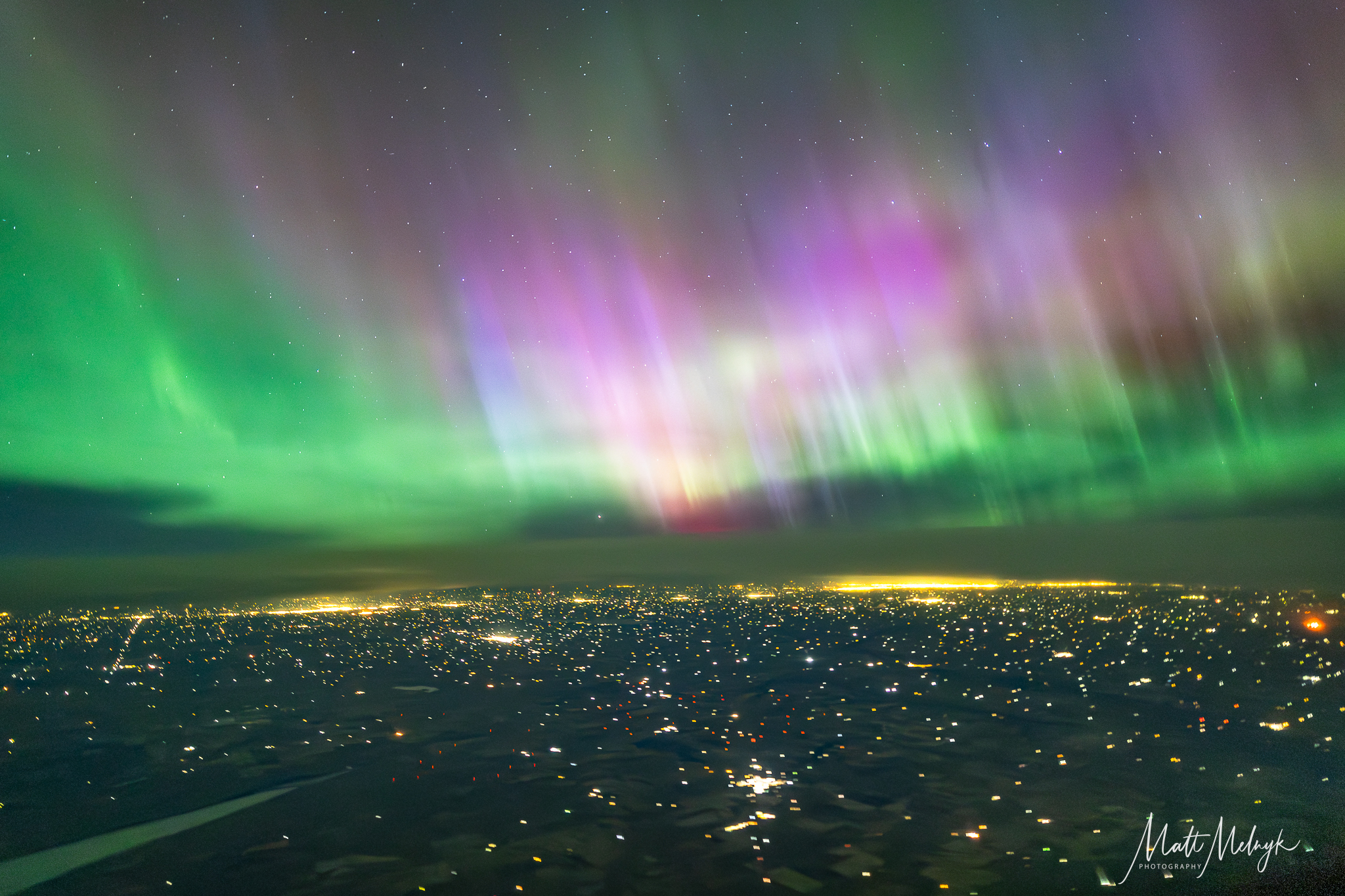 curtains of magenta and green light dance in the sky, yellow spots from city lights fill the scene below. 