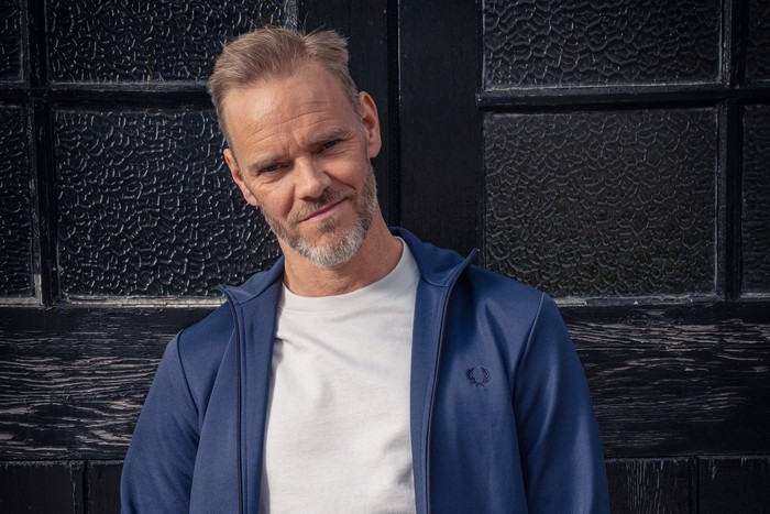 Ray (Joe Absolom) stands against doors in a blue jacket and white t-shirt