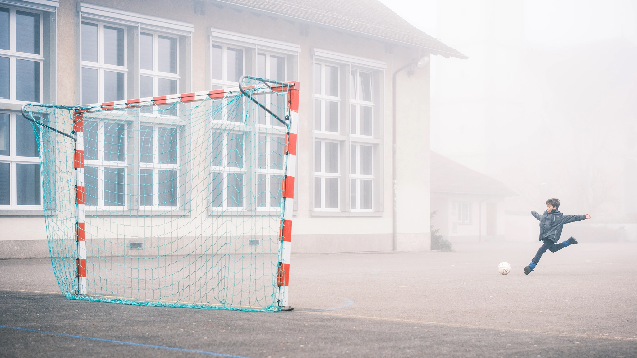 A child in a blue jacket prepares to kick a soccer ball towards a net on a foggy playground, with school building windows in the background