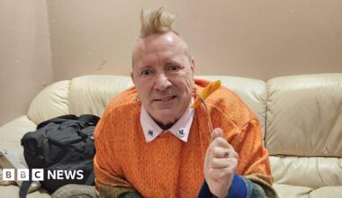 An older man, John Lydon, sitting on a cream leather sofa, he is wearing an orange jumper and holding up a fork with an orange chip on. He has a blonde mohawk and is looking into the camera.