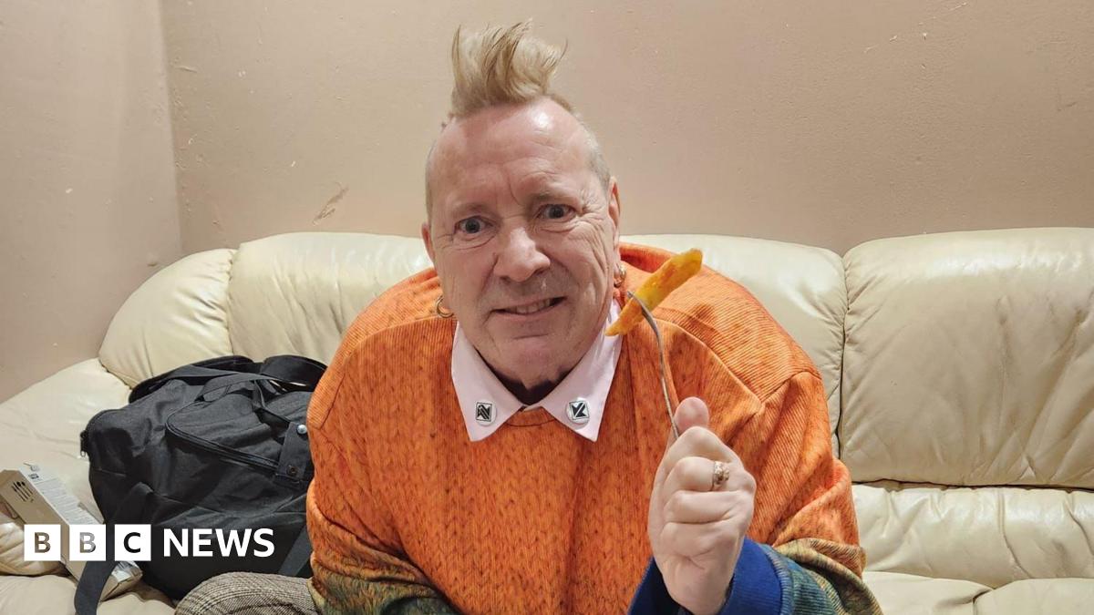 An older man, John Lydon, sitting on a cream leather sofa, he is wearing an orange jumper and holding up a fork with an orange chip on. He has a blonde mohawk and is looking into the camera.