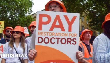 A medical worker in scrubs is pictured in a hospital ward leaning forward and taking a tray from a table. A patient on the other side of the table is obscured by a curtain, with just their hand visible. They are holding a tissue.