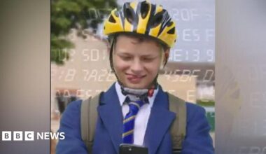 A boy in a blue blazer and striped tie has a yellow helmet on and a rucksack, and he is looking down at a phone. He is sticking his tongue out to one side of his mouth. He is outside.