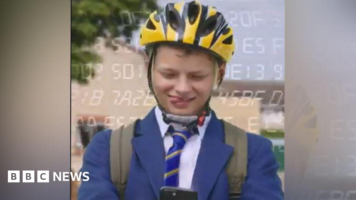 A boy in a blue blazer and striped tie has a yellow helmet on and a rucksack, and he is looking down at a phone. He is sticking his tongue out to one side of his mouth. He is outside.