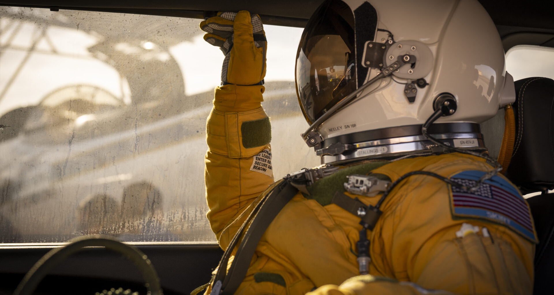 A person facing left sits inside a vehicle. They are wearing a yellow jumpsuit and gloves and a white helmet with a visor that looks like a smaller version of the kind astronauts wear during spacewalks. Outside of the window behind the person, you can see the ER-2 aircraft being prepared for flight.