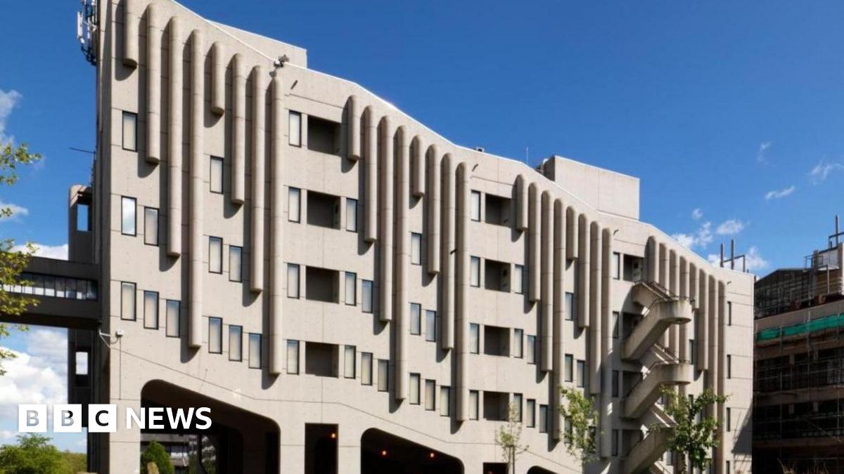 A large, grey-coloured, modern concrete building with an unusual architectural design. There are four storeys with black-rimmed windows. In the background the sky is blue scattered with white clouds. To the right of the building is another building with several storeys.