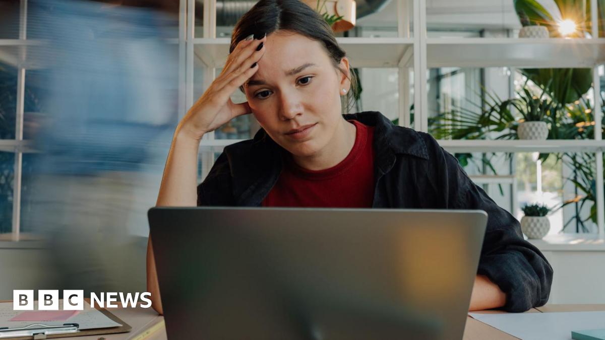 A young woman wearing a dark blue shirt and a red t-shirt looks concerned as she looks at her laptop. She has her right hand on the side of her head.
