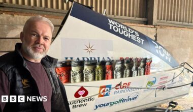 Ollie Phillips smiling at the camera is wearing a burgundy jumper and black gilet with a rowing boat in the background.