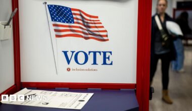 A blue table is blocked off by a white and red VOTE privacy screen. There is a ballot lying on the table and there appear to be people in the background in rack focus.