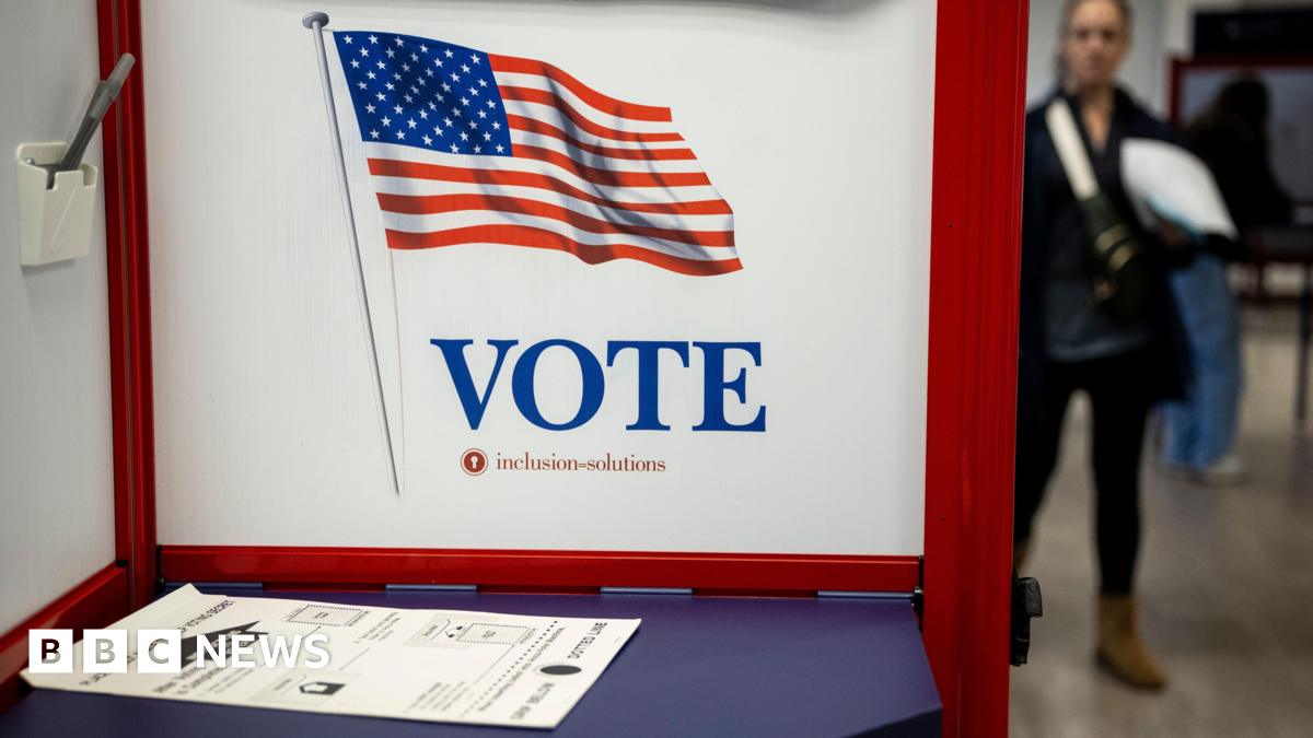 A blue table is blocked off by a white and red VOTE privacy screen. There is a ballot lying on the table and there appear to be people in the background in rack focus.