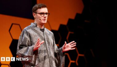 Welsh computer scientist Llion jones stands on stage at a TED-AI conference in San Francisco, wearing a grey patterned, loose-fitting shirt. The background features a bold orange shape with a honeycomb-style black geometric design. Llion wears glasses and a microphone and is mid-presentation, with both hands raised in a gesturing position.
