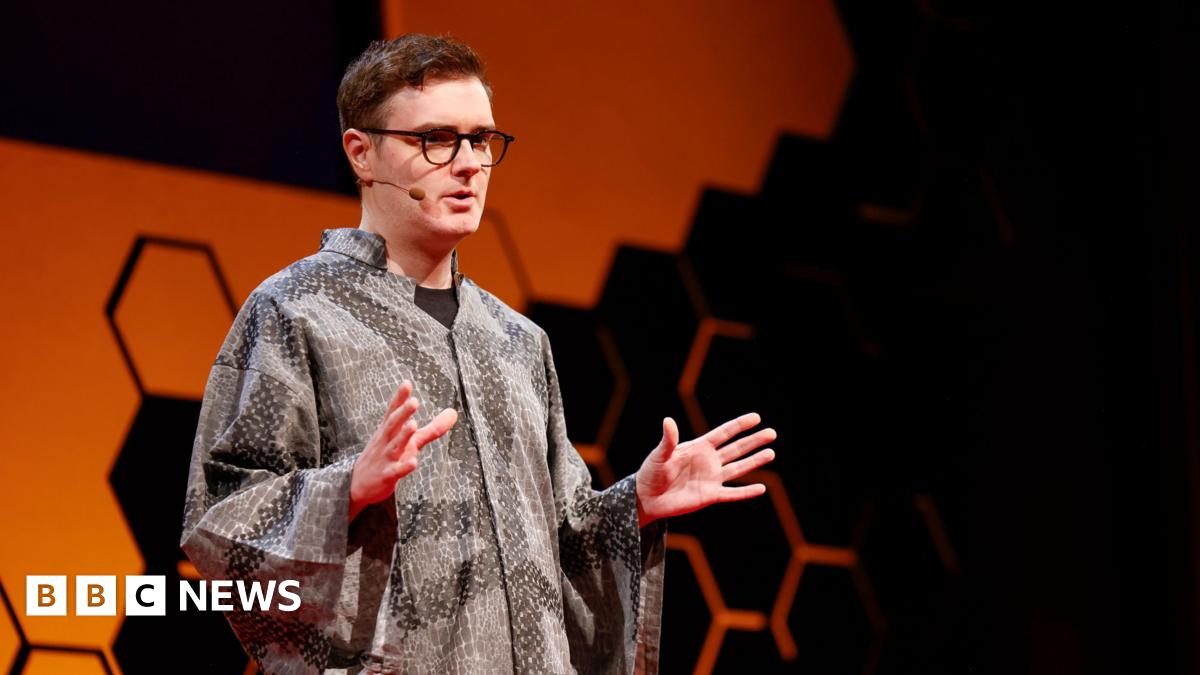 Welsh computer scientist Llion jones stands on stage at a TED-AI conference in San Francisco, wearing a grey patterned, loose-fitting shirt. The background features a bold orange shape with a honeycomb-style black geometric design. Llion wears glasses and a microphone and is mid-presentation, with both hands raised in a gesturing position.