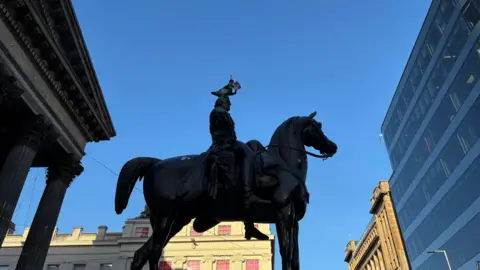A statue of a man sitting atop a horse. A second statue, of a pigeon, has been added to his head. It is a sunny and bright day.