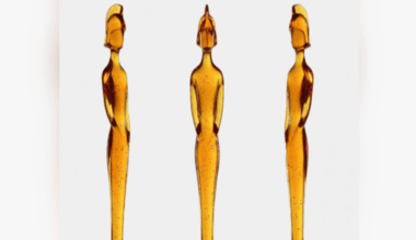 Three identical statues are displayed side by side against a white background. Each statue is made of translucent amber-colored material with small dark speckles throughout. The statues have an elongated, elegant form resembling a stylized human figure standing upright with arms close to the body. The base of each statue is a rounded, bulb-like shape that gradually narrows into a tall, slender torso and head. The figures have a helmet-like headpiece, giving them a distinctive silhouette.