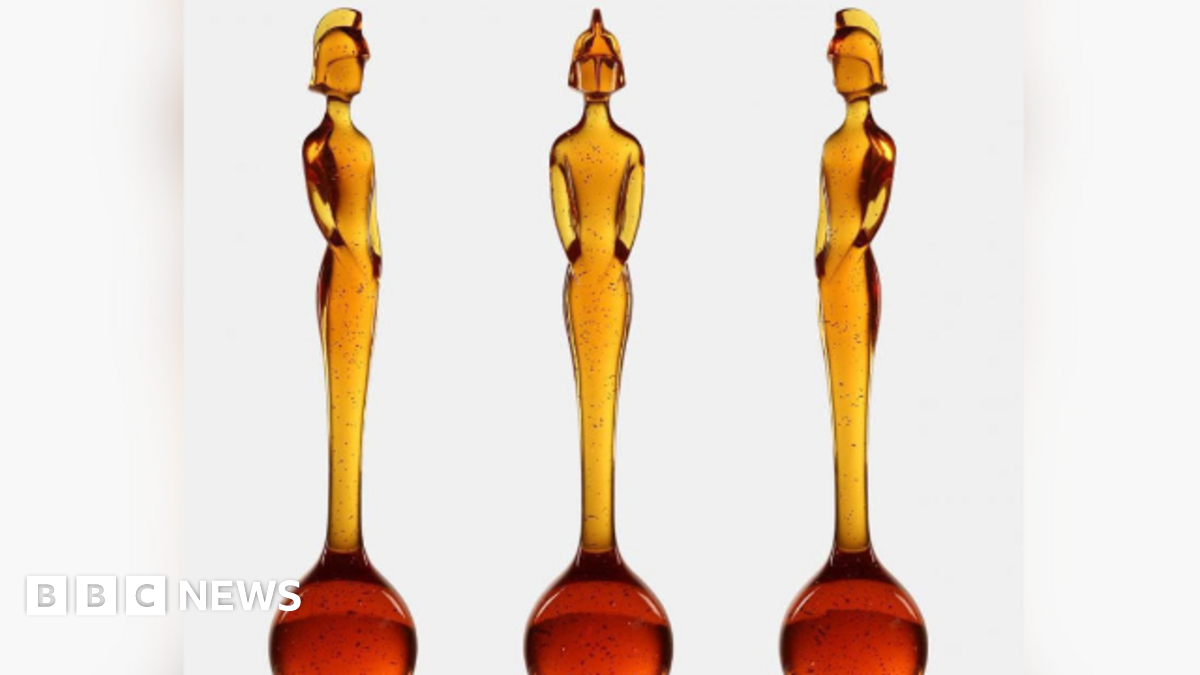 Three identical statues are displayed side by side against a white background. Each statue is made of translucent amber-colored material with small dark speckles throughout. The statues have an elongated, elegant form resembling a stylized human figure standing upright with arms close to the body. The base of each statue is a rounded, bulb-like shape that gradually narrows into a tall, slender torso and head. The figures have a helmet-like headpiece, giving them a distinctive silhouette.