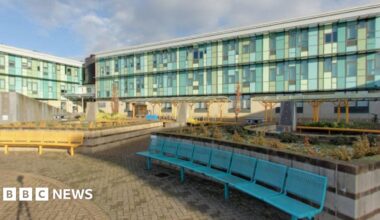 A screengrab taken of the outside of St Brendan's Sixth Form College. The Image shows a big school campus with light blue glass windows. There are some flower beds in the middle of the courtyard with long yellow and blue benches placed in front of them.