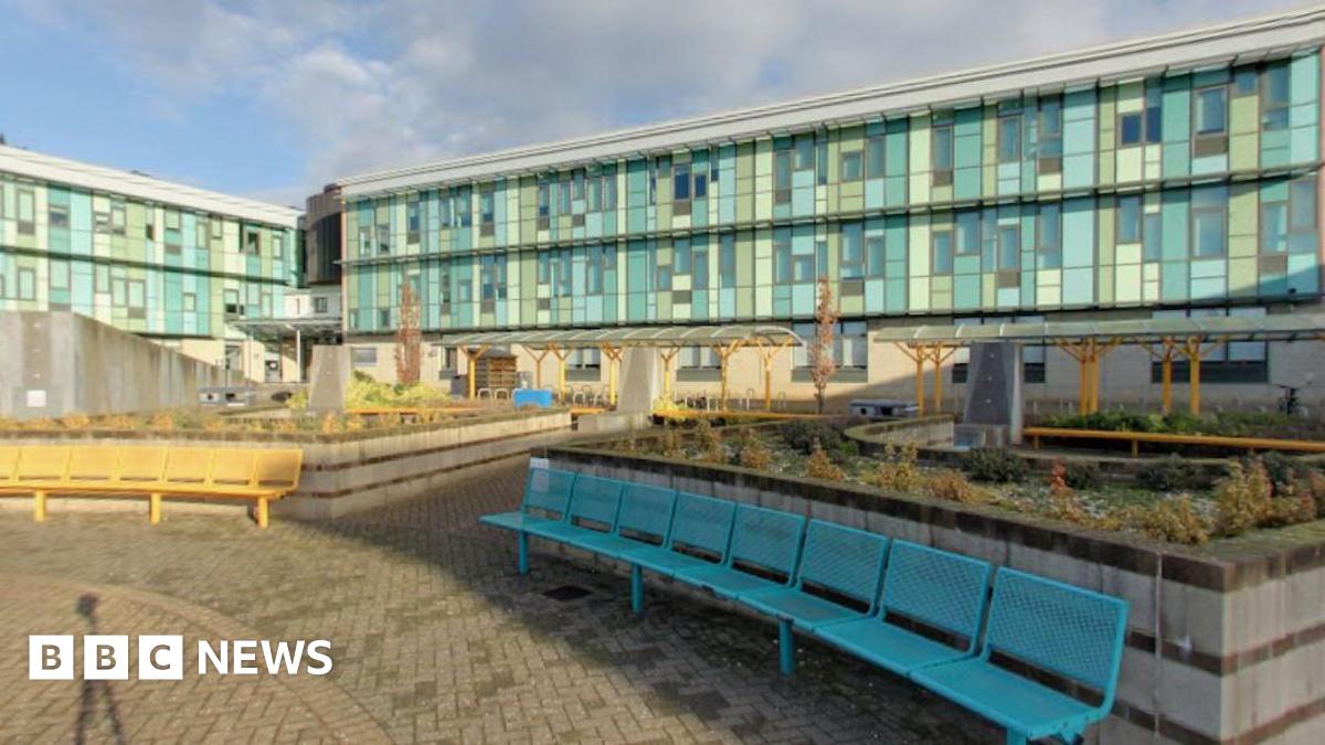 A screengrab taken of the outside of St Brendan's Sixth Form College. The Image shows a big school campus with light blue glass windows. There are some flower beds in the middle of the courtyard with long yellow and blue benches placed in front of them.