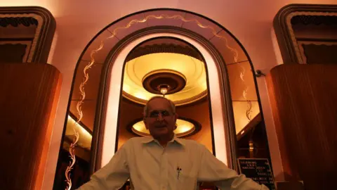 Getty Images Nazir Hoosein, owner of Liberty Cinema, pictured smiling into the camera. His figure is framed by an arch, with circular Art Deco lights also behind him.