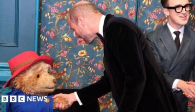The Prince of Wales, wearing a black tuxedo, shaking the hand of an actor in costume as Paddington Bear - Tom Fletcher from McFly can be seen in the background