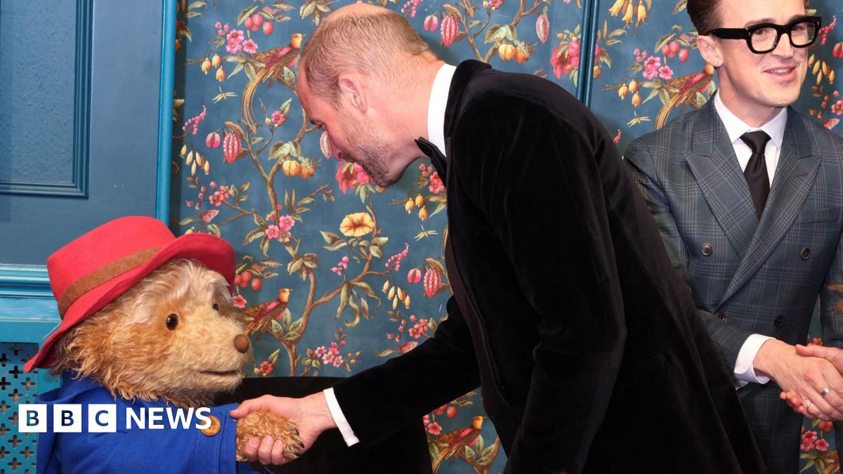 The Prince of Wales, wearing a black tuxedo, shaking the hand of an actor in costume as Paddington Bear - Tom Fletcher from McFly can be seen in the background
