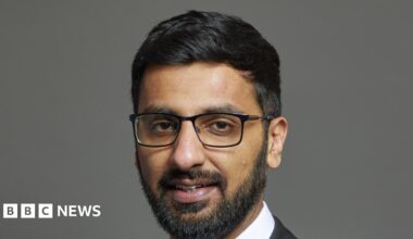 Official portrait of Adnan Hussain taken against a grey backdrop.  He has black hair and a beard and is wearing glasses.