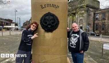 A couple in Motorhead themed T-shirts stand either side of the plinth of the Lemmy statue, which has Lemmy engraved on it. Also on the plinth is is Motorhead's large black motif, which is known as the Snaggletooth or the War Pig and sometimes the Iron Boar. The statue is in a square, with roads and buildings surrounding it.