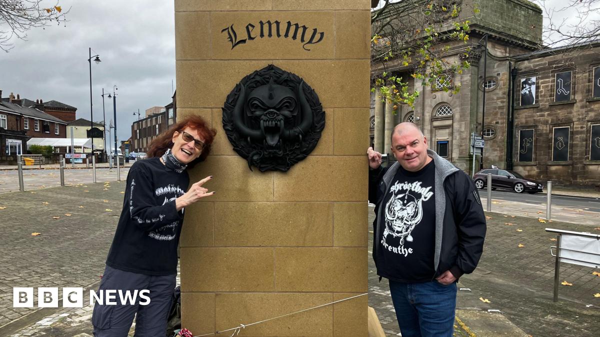 A couple in Motorhead themed T-shirts stand either side of the plinth of the Lemmy statue, which has Lemmy engraved on it. Also on the plinth is is Motorhead's large black motif, which is known as the Snaggletooth or the War Pig and sometimes the Iron Boar. The statue is in a square, with roads and buildings surrounding it.