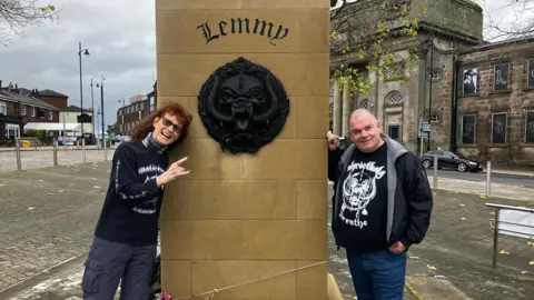 BBC A couple in Motorhead themed T-shirts stand either side of the plinth of the Lemmy statue, which has Lemmy engraved on it. Also on the plinth is is Motorhead's large black motif, which is known as the Snaggletooth or the War Pig and sometimes the Iron Boar. The statue is in a square, with roads and buildings surrounding it.