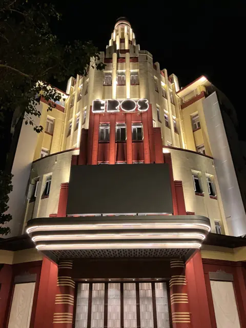Art Deco Mumbai Trust A night-time image of a lit up Eros Cinema, a Unesco-designated Art Deco theatre at Churchgate in Mumbai. The multi-storey building, which has a spherical tower at its top, is painted cream and partially faced with red sandstone. 
