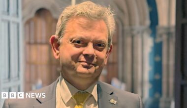 Anton Muscatelli stands inside Glasgow University. The background is the Gothic architecture of the university's main buidling. He is framed by two this wooden doors. Sir Anton wears a grey jacket with a white shirt and yellow tie.