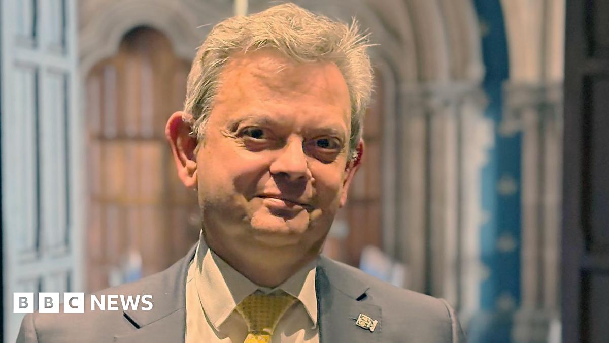 Anton Muscatelli stands inside Glasgow University. The background is the Gothic architecture of the university's main buidling. He is framed by two this wooden doors. Sir Anton wears a grey jacket with a white shirt and yellow tie.