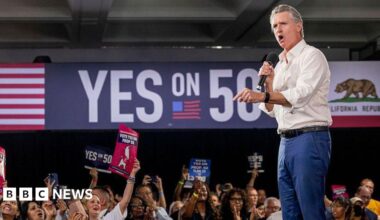 California Gov Gavin Newsom appears on stage in support of a redistricting effort in his state. People in the audience are holding up signs in support of Prop 50 and on the wall in the background is a "YES ON 50" sign.