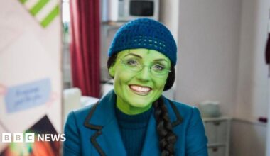 Kerry Ellis sits in a dress room in a chair and smiles at the camera. She has green face paint on to match her character Elphaba. She wears a blue blazer, blue jumper underneath and a blue hat. She is also wearing a black wig that has been braided.