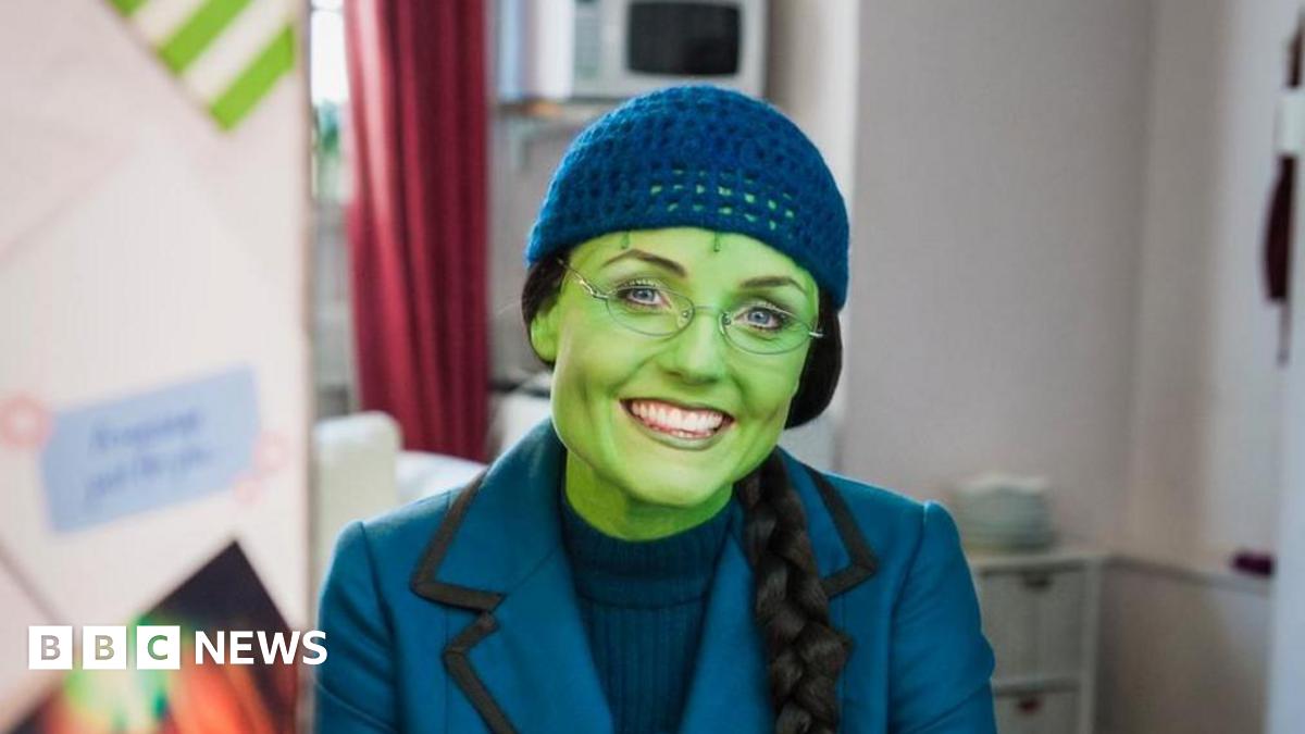 Kerry Ellis sits in a dress room in a chair and smiles at the camera. She has green face paint on to match her character Elphaba. She wears a blue blazer, blue jumper underneath and a blue hat. She is also wearing a black wig that has been braided.