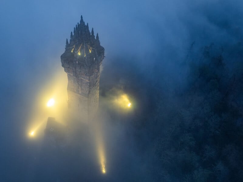 A tall, illuminated stone tower rises above dense fog at night, with spotlights shining through the mist and casting a mysterious glow on the surrounding dark landscape.