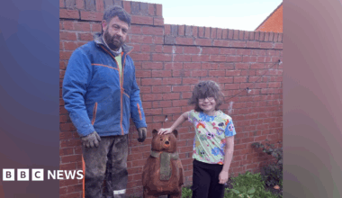 A young girl with short wavy brown hair is wearing glasses and a multi-coloured T-shirt and has her hand on top of a small brown wooden bear with green scarf sculpture in a garden. A man stands on the other side of the bear. He has brown hair and a beard, and is wearing a blue waterproof jacket and black trousers with sawdust on them, as well as black gloves.