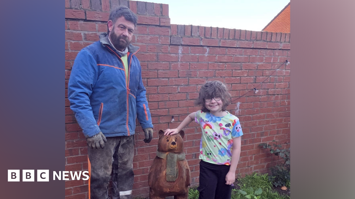A young girl with short wavy brown hair is wearing glasses and a multi-coloured T-shirt and has her hand on top of a small brown wooden bear with green scarf sculpture in a garden. A man stands on the other side of the bear. He has brown hair and a beard, and is wearing a blue waterproof jacket and black trousers with sawdust on them, as well as black gloves.
