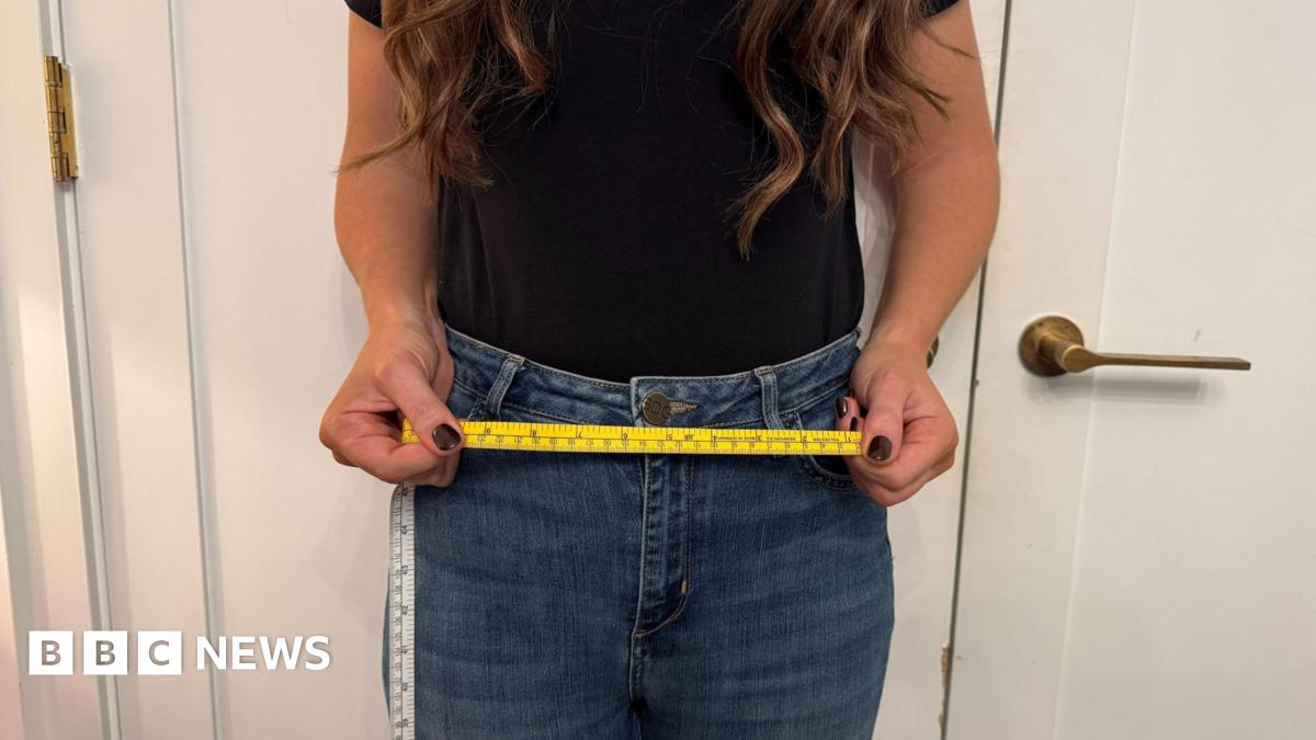 A woman in jeans stands with a yellow tape measure across her waist.