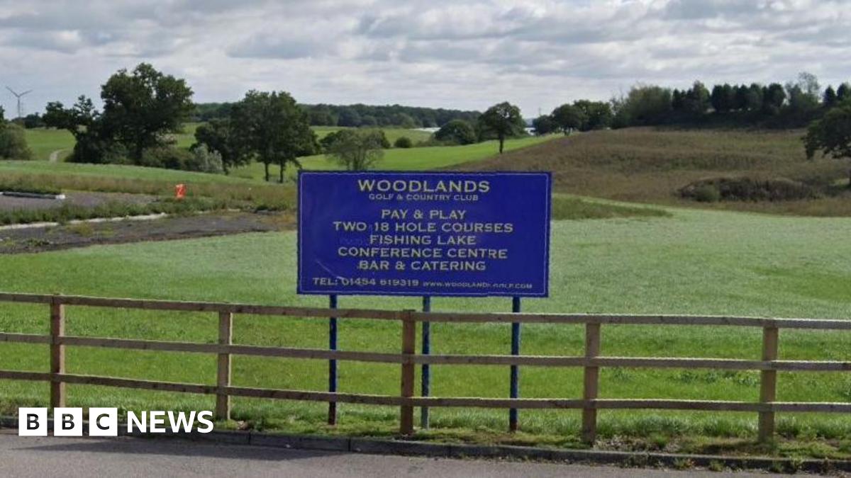 A blue sign saying Woodlands Golf and Country Club, above a fence with green grass and trees behind.