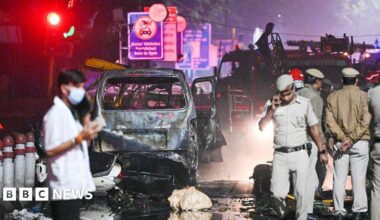Police officers and officials walk around the nighttime scene of a deadly car blast in India's capital city, Delhi. Behind them is the charred wreck of a burnt out car and a fire engine. The road is wet from firefighting efforts and covered in debris.