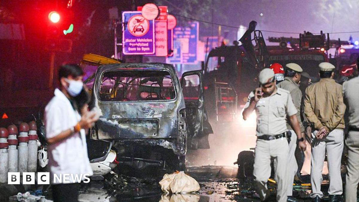 Police officers and officials walk around the nighttime scene of a deadly car blast in India's capital city, Delhi. Behind them is the charred wreck of a burnt out car and a fire engine. The road is wet from firefighting efforts and covered in debris.