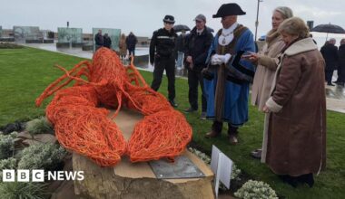 The sculpture is srrounded by a group of people who are examining it.  It sits on a stone plinth surrounded by planting and the wire lobst is bright orange.