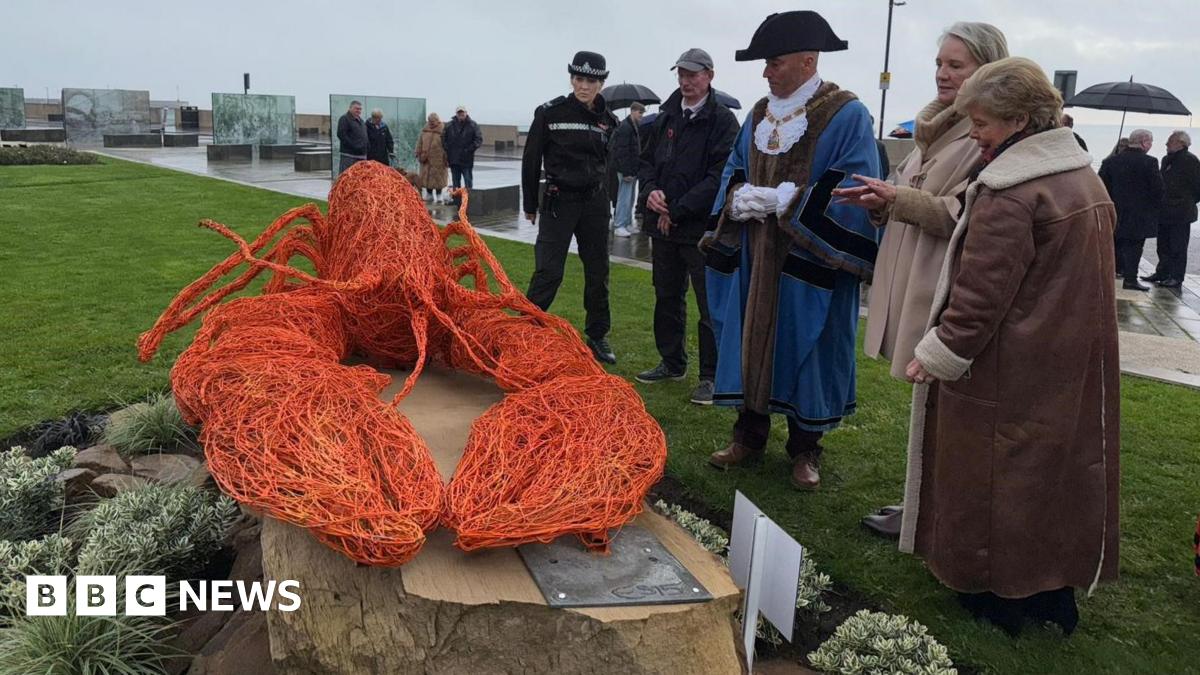 The sculpture is srrounded by a group of people who are examining it.  It sits on a stone plinth surrounded by planting and the wire lobst is bright orange.