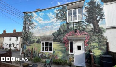 Huge mural painted on a house. She is stood near to the house's white door. The wall is painted with grass, lots of colourful flowers, butterflies and there is a countryside landscape.