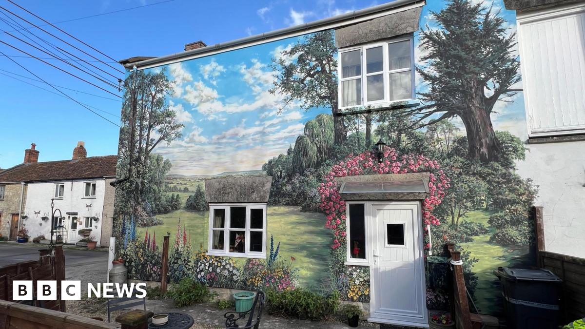 Huge mural painted on a house. She is stood near to the house's white door. The wall is painted with grass, lots of colourful flowers, butterflies and there is a countryside landscape.